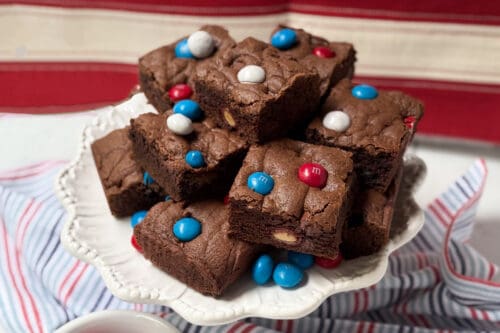 A plate of chocolate M&M cookie bars topped with red, white, and blue candies is displayed on a white cake stand with a striped cloth and a red, white, and beige backdrop.