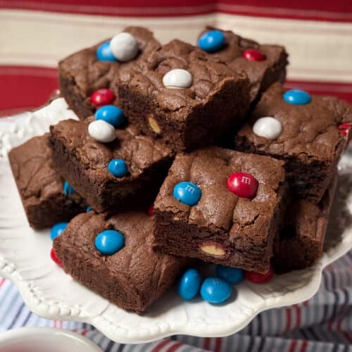 A plate of chocolate M&M cookie bars topped with red, white, and blue candies is displayed on a white cake stand with a striped cloth and a red, white, and beige backdrop.