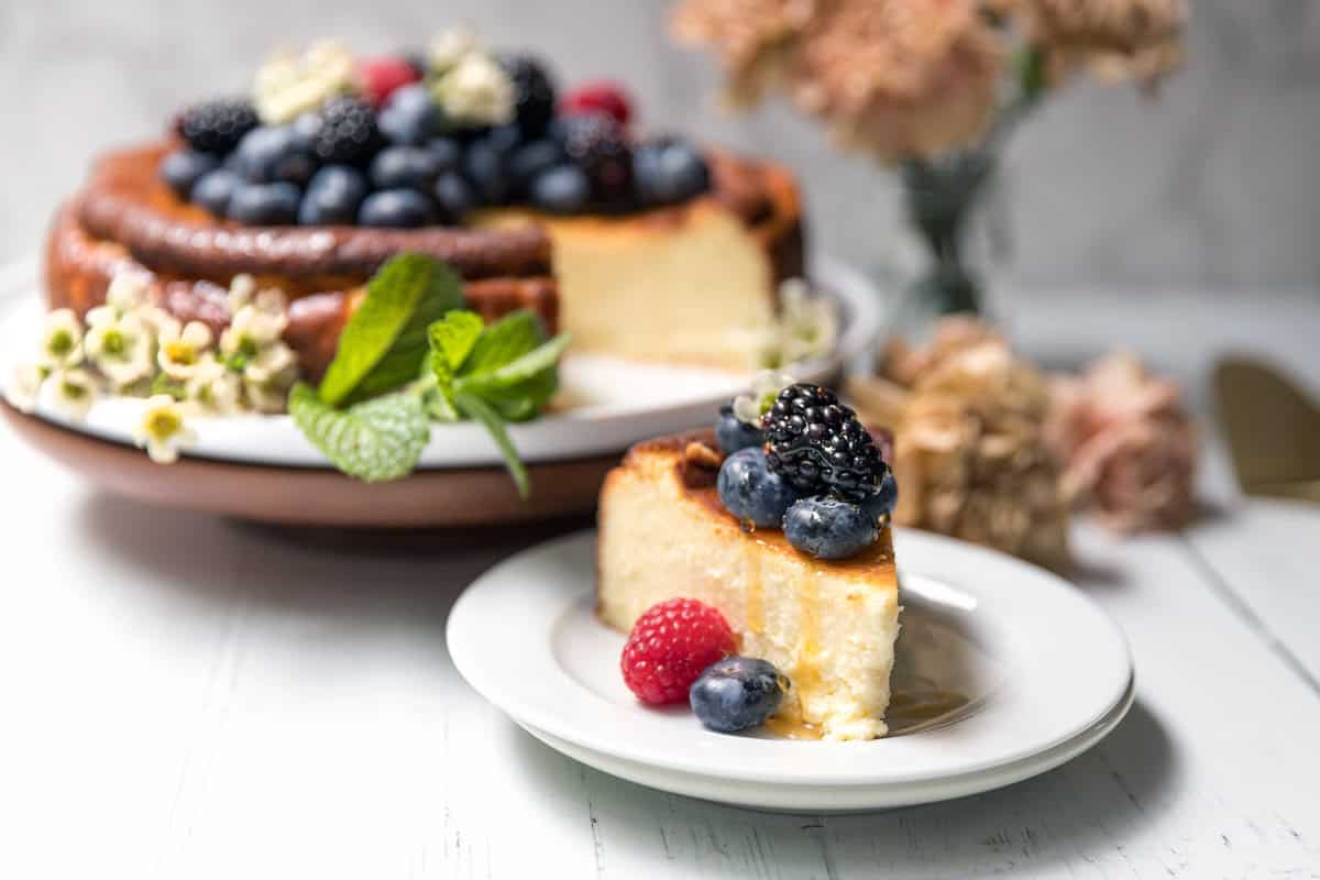 A slice of basque cheesecake topped with fresh berries and honey sits on a plate in the foreground, with the rest of the cheesecake and more berries in the background, surrounded by flowers and mint leaves.