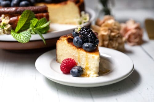 A slice of basque cheesecake topped with blueberries, a blackberry, and a raspberry sits on a white plate. The rest of the cheesecake, garnished with mint, is in the background on a cake stand.