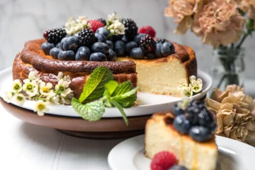 A basque cheesecake topped with blueberries, blackberries, raspberries, and small white flowers sits on a plate, with a slice served on a smaller plate nearby. A vase of light brown carnations is in the background.
