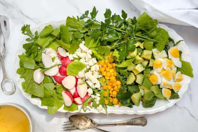 A platter of salad with leafy greens, sliced radishes, crumbled feta, corn, diced avocado, chopped hard-boiled eggs, and parsley, arranged in rows on a white tray. Silver utensils and a bowl of dressing are nearby.