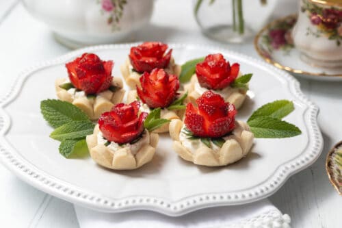 A white plate displays six strawberry tarts topped with strawberries carved into rose shapes and garnished with fresh mint leaves. The background includes floral teacups and a teapot.