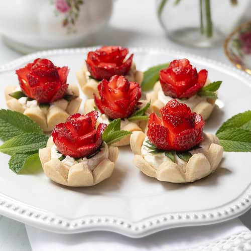 A white plate displays six strawberry tarts topped with strawberries carved into rose shapes and garnished with fresh mint leaves. The background includes floral teacups and a teapot.