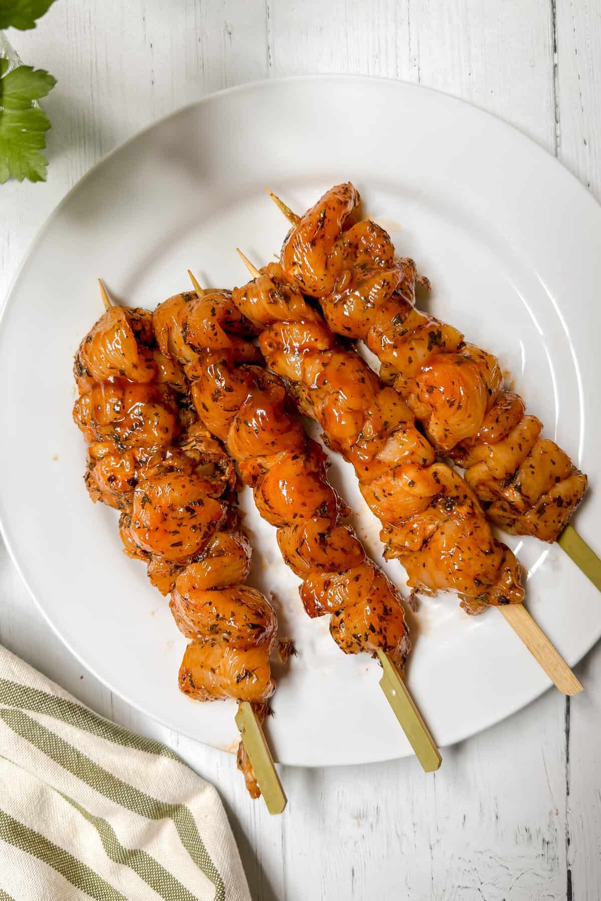 Four skewers of marinated raw chicken are arranged on a white plate atop a white wooden surface, with a striped towel and green herbs partially visible at the edges.