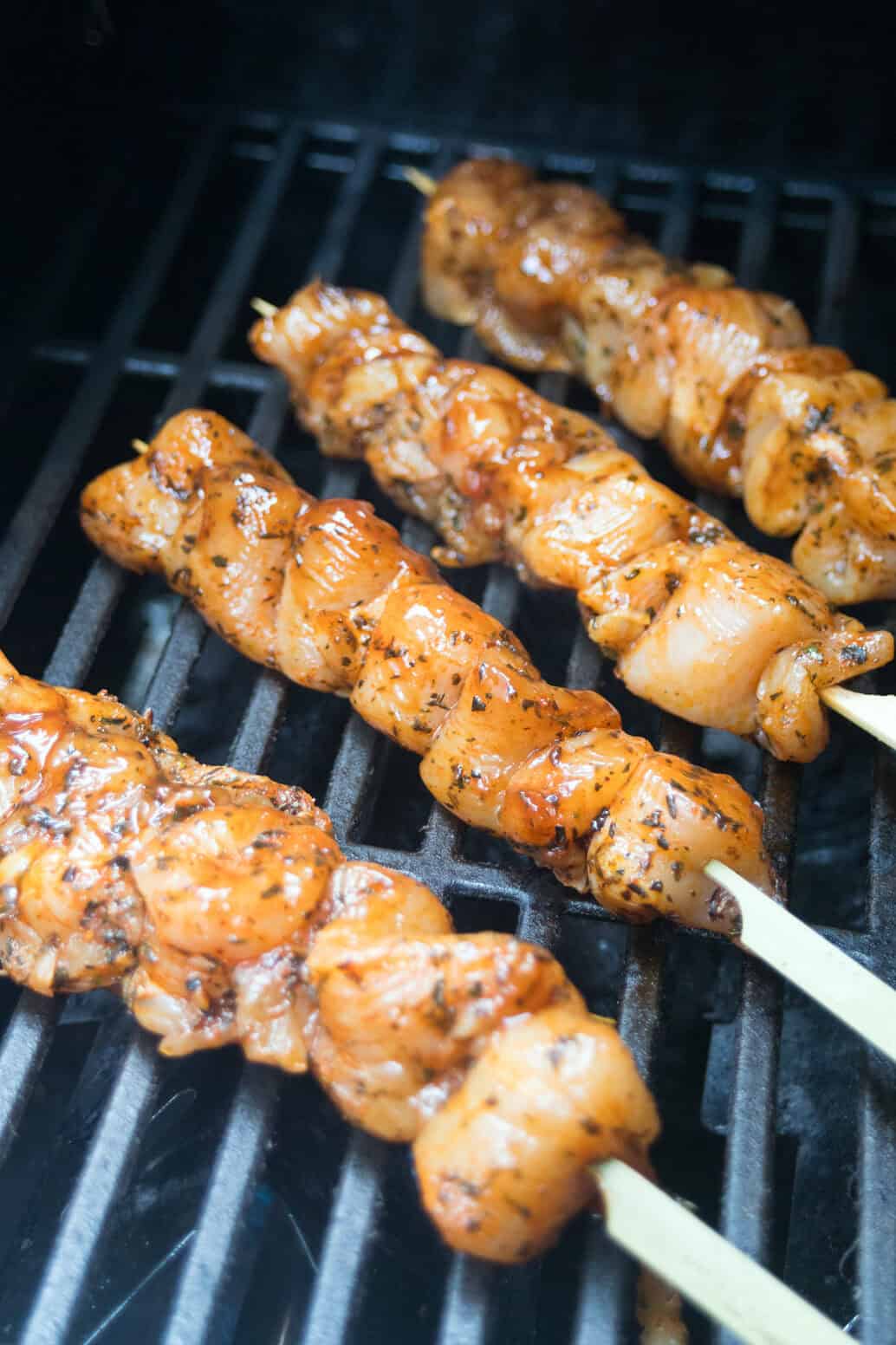 Four skewers of marinated chicken are grilling on a barbecue, with the meat sizzling and slightly charred, set against the dark grill grates.