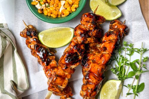 Three grilled chicken skewers with barbecue sauce are served on parchment with lime wedges, fresh herbs, a bowl of corn topped with cheese, and a striped napkin on a wooden tray.