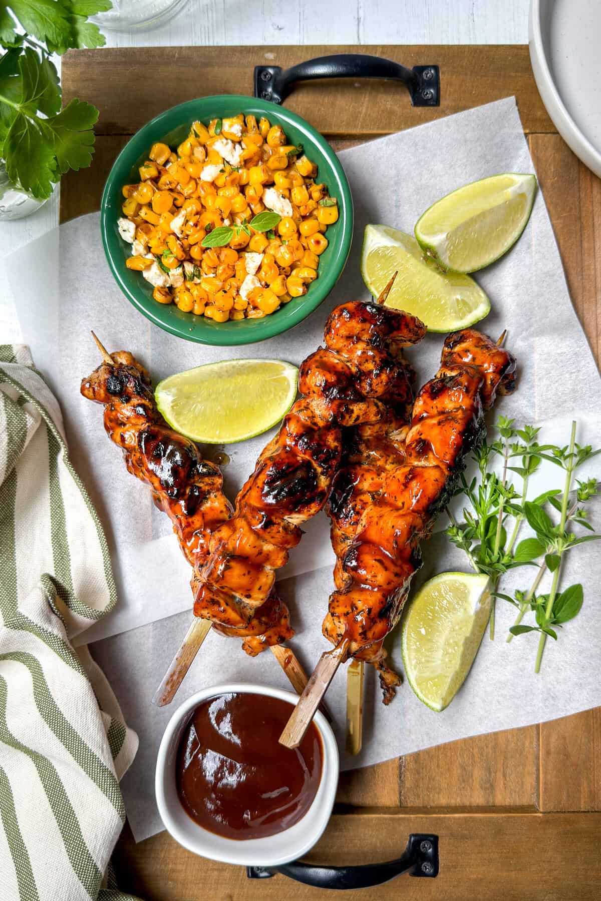 Three grilled chicken skewers with barbecue sauce are served on parchment with lime wedges, fresh herbs, a bowl of corn topped with cheese, and a striped napkin on a wooden tray.