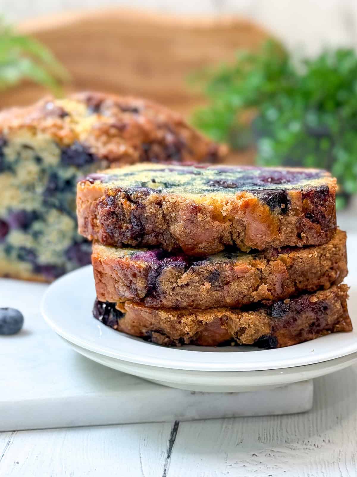 Three slices of blueberry muffin bread stacked on a white plate, with more blueberry bread and a green plant in the blurred background.