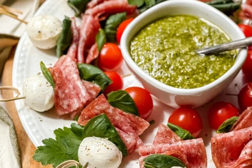 A white plate with skewers of salami, mozzarella balls, cherry tomatoes, and basil, arranged in a circle around a bowl of green pesto dip. The plate sits on a wooden board.