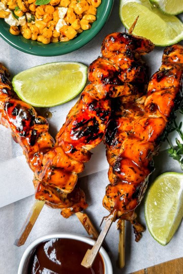 Three BBQ chicken skewers with barbecue sauce, lime wedges, grilled corn, and fresh herbs on parchment paper.