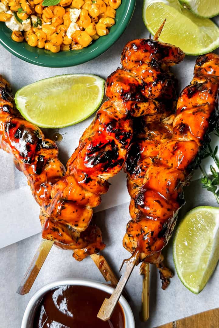 Three BBQ chicken skewers with barbecue sauce, lime wedges, grilled corn, and fresh herbs on parchment paper.