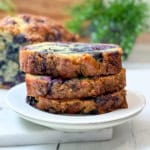 Three slices of blueberry muffin bread loaf cake are stacked on a white plate, with a blurred background showing more cake and green foliage.