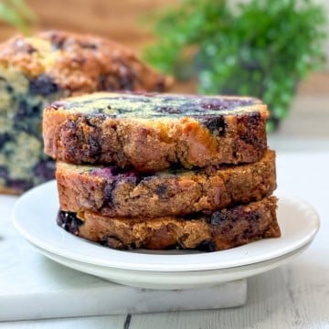 Three slices of blueberry muffin bread loaf cake are stacked on a white plate, with a blurred background showing more cake and green foliage.