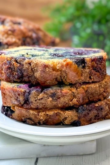 Three slices of blueberry muffin bread loaf cake are stacked on a white plate, with a blurred background showing more cake and green foliage.