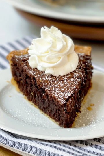 A slice of chocolate pie topped with whipped cream and powdered sugar on a white plate, set on a striped cloth—perfect for Father's Day dessert recipes.