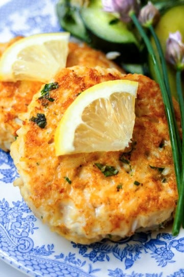 Two Maryland crab cakes topped with lemon wedges, served with corn on the cob and a cucumber salad on a blue and white plate.