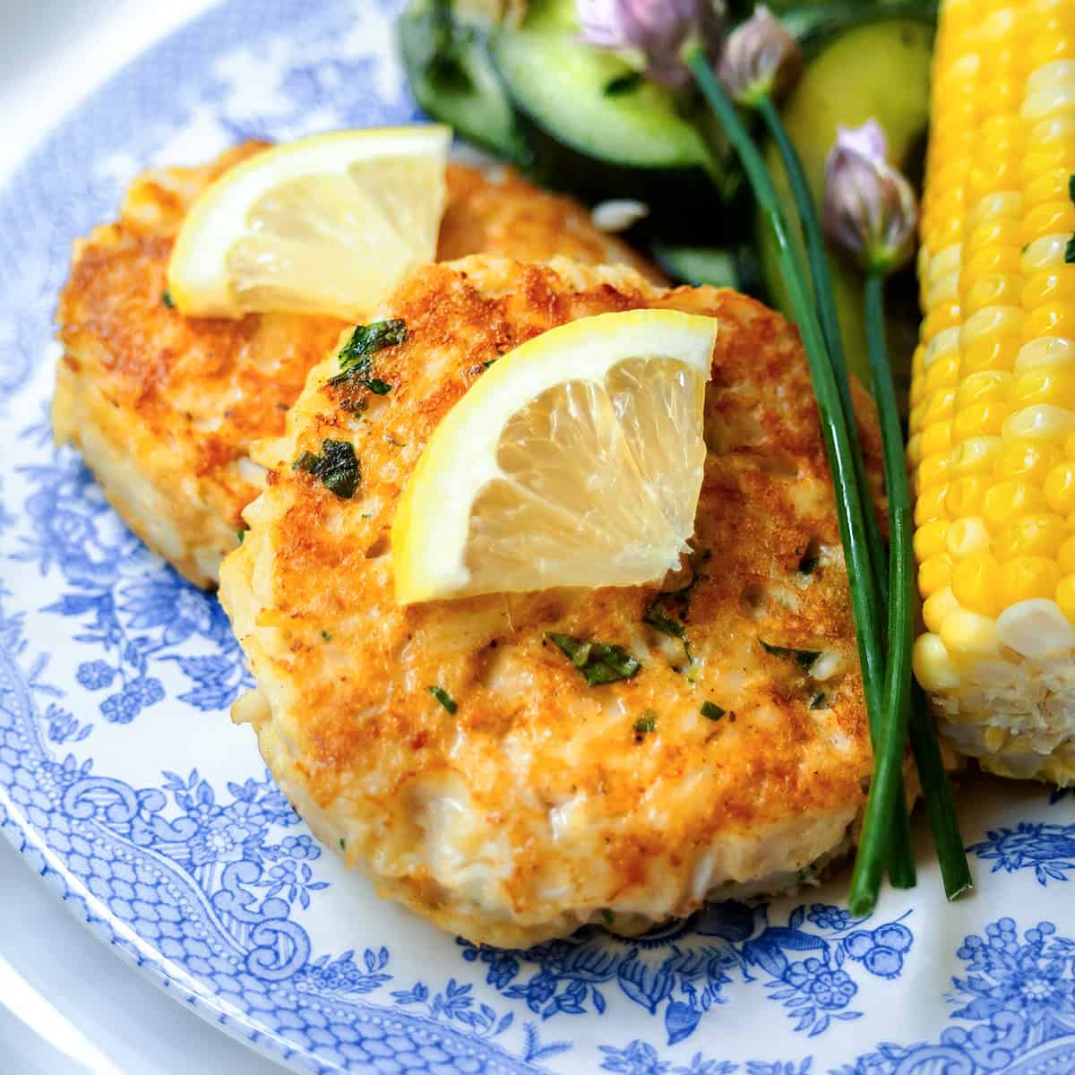 Two Maryland crab cakes topped with lemon wedges, served with corn on the cob and a cucumber salad on a blue and white plate.
