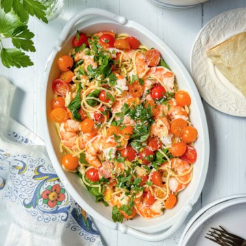Oval dish with spaghetti, shrimp, cherry tomatoes, parsley, and grated cheese-this shrimp pasta recipe sits on a white table with napkin, plate, and cutlery nearby.
