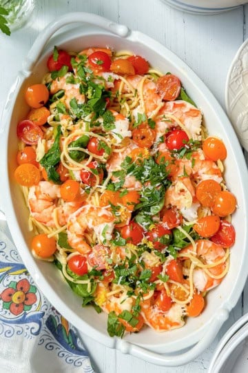Oval dish with spaghetti, shrimp, cherry tomatoes, parsley, and grated cheese—this shrimp pasta recipe sits on a white table with napkin, plate, and cutlery nearby.