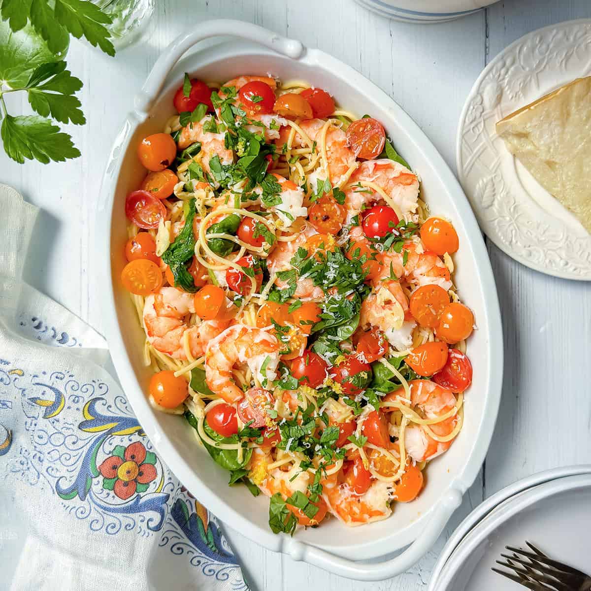 Oval dish with spaghetti, shrimp, cherry tomatoes, parsley, and grated cheese-this shrimp pasta recipe sits on a white table with napkin, plate, and cutlery nearby.
