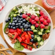 A bowl of spinach strawberry salad with blueberries, raspberries, cucumber, pecans, and crumbled feta cheese sits beside a jar of dressing.