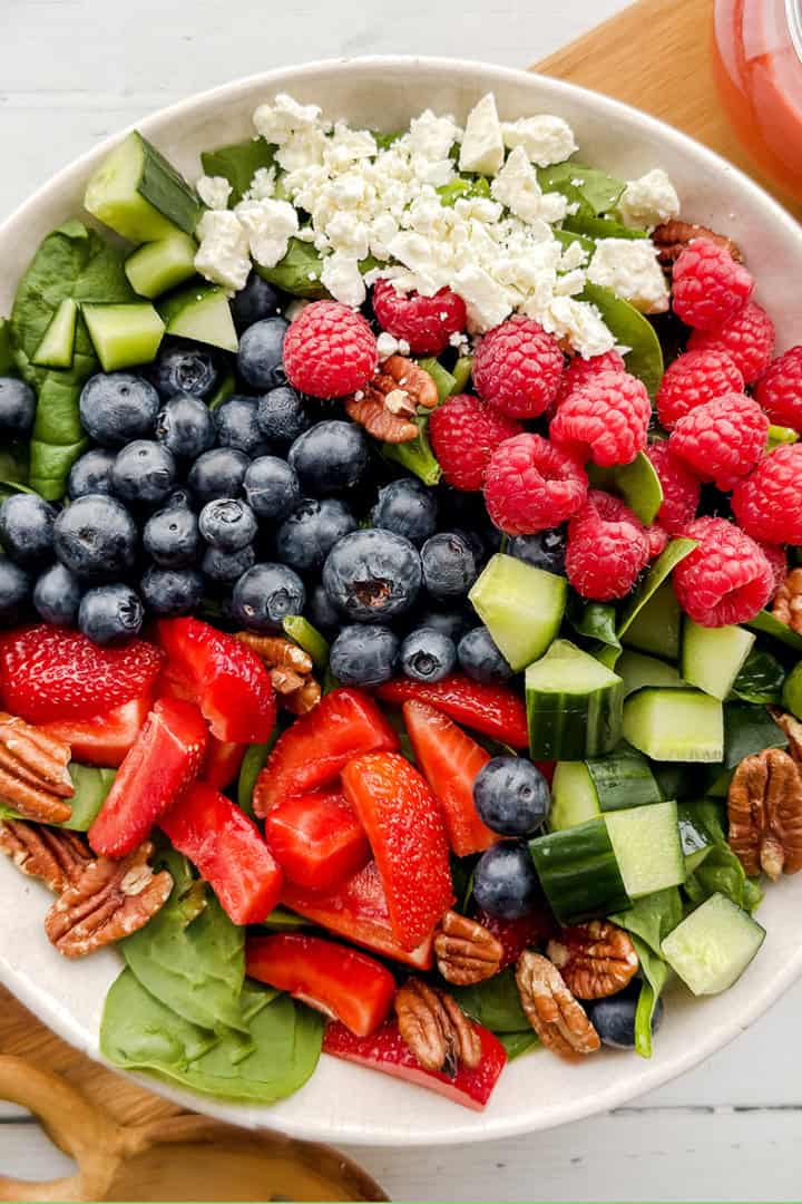 A bowl of spinach strawberry salad with blueberries, raspberries, cucumber, pecans, and crumbled feta cheese sits beside a jar of dressing.
