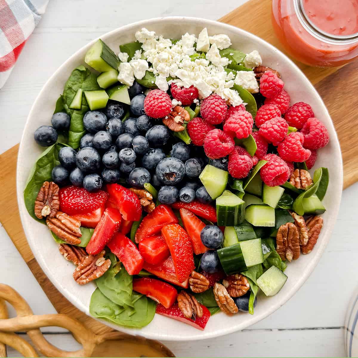 A bowl of spinach strawberry salad with blueberries, raspberries, cucumber, pecans, and crumbled feta cheese sits beside a jar of dressing.