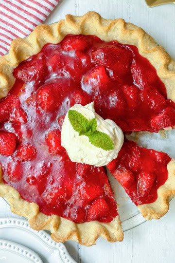A strawberry pie with a slice missing, topped with whipped cream and mint, sits on a white wooden table next to a striped cloth and plates—perfect for trying your favorite strawberry pie recipe.