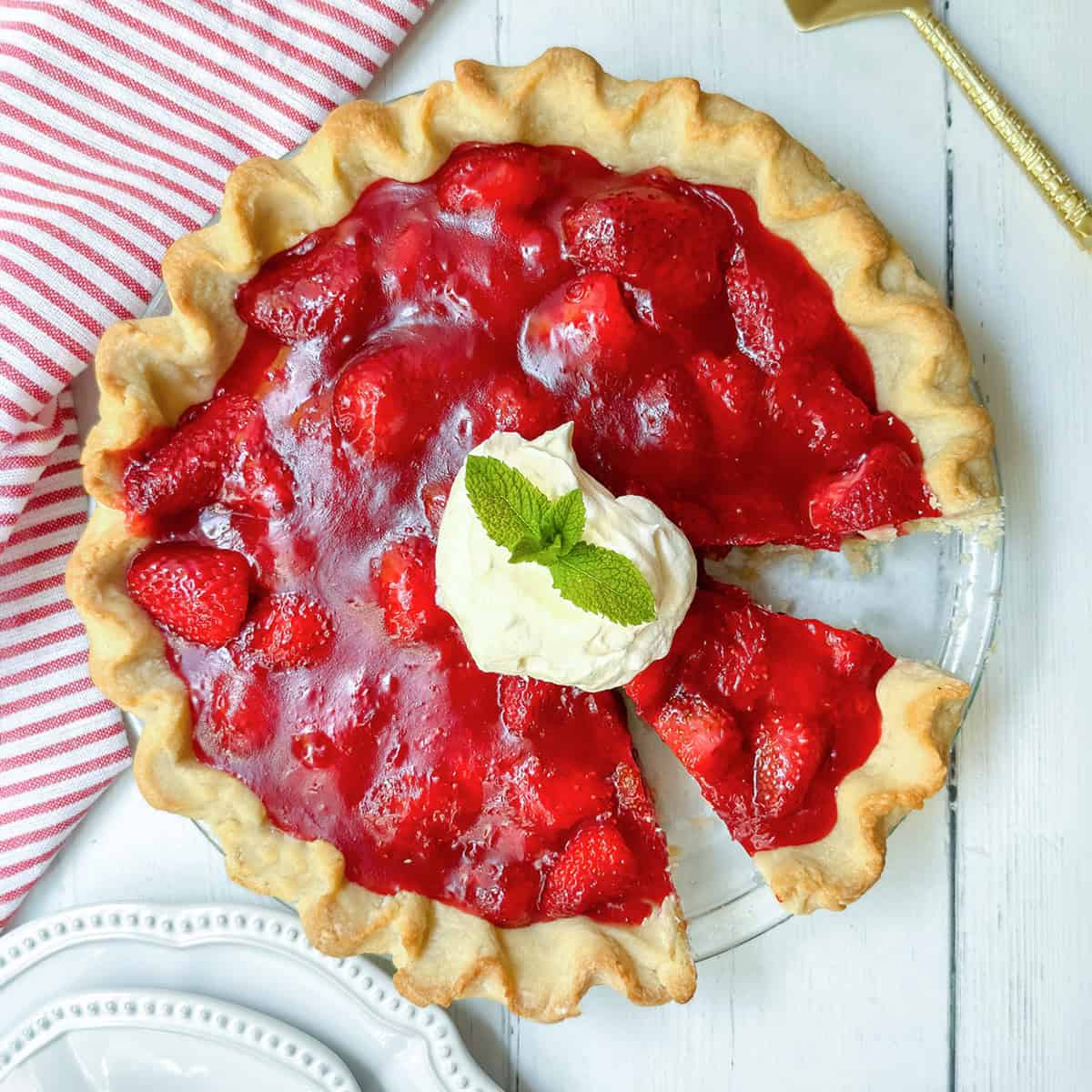 A strawberry pie with a slice missing, topped with whipped cream and mint, sits on a white wooden table next to a striped cloth and plates—perfect for trying your favorite strawberry pie recipe.