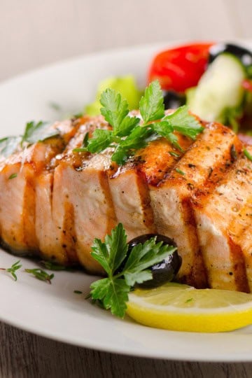 Honey glazed salmon fillet garnished with parsley and served with lemon slices, black olives, and a side salad of lettuce, tomato, and cucumber on a white plate.
