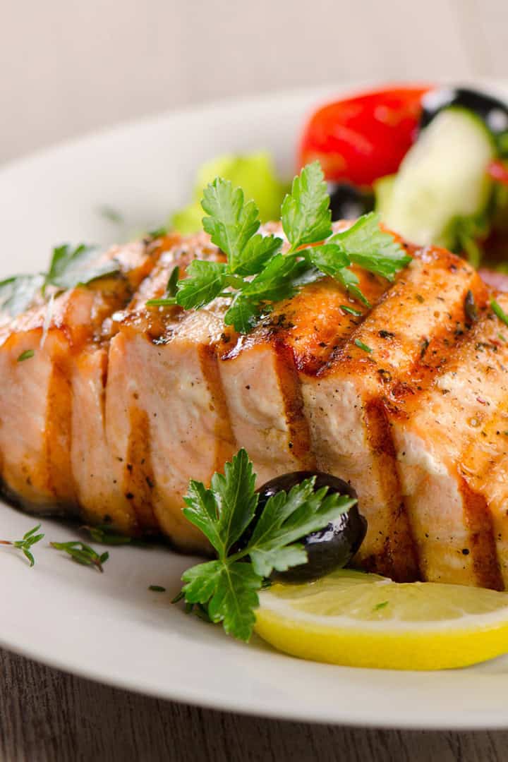 Honey glazed salmon fillet garnished with parsley and served with lemon slices, black olives, and a side salad of lettuce, tomato, and cucumber on a white plate.
