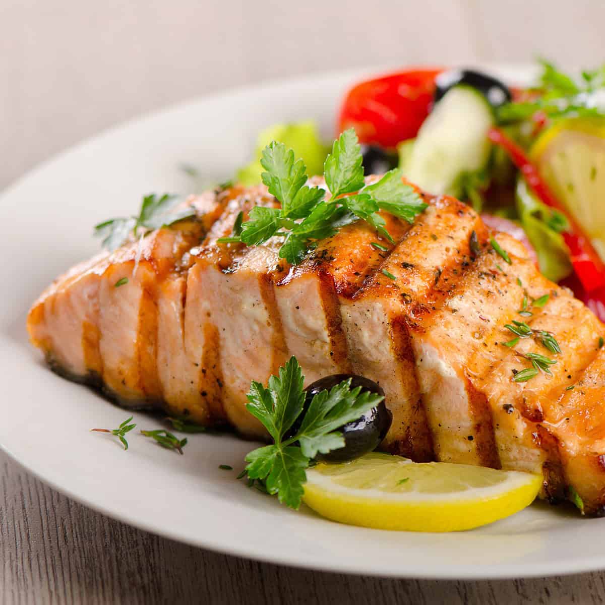 Honey glazed salmon fillet garnished with parsley and served with lemon slices, black olives, and a side salad of lettuce, tomato, and cucumber on a white plate.