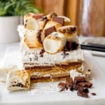 A no-bake s'mores icebox cake sits on a white surface, layered and topped with toasted marshmallows and chocolate pieces, with extra chocolate and marshmallows scattered in front.