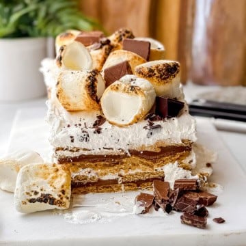 A no-bake s'mores icebox cake sits on a white surface, layered and topped with toasted marshmallows and chocolate pieces, with extra chocolate and marshmallows scattered in front.