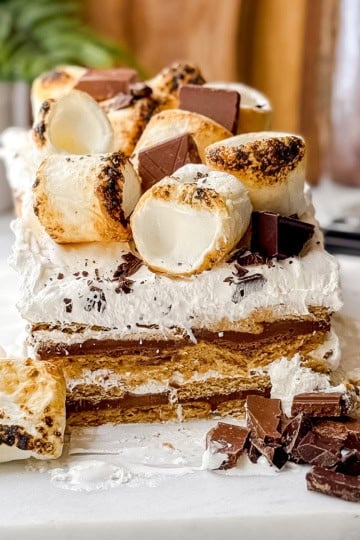 A no-bake s'mores icebox cake sits on a white surface, layered and topped with toasted marshmallows and chocolate pieces, with extra chocolate and marshmallows scattered in front.