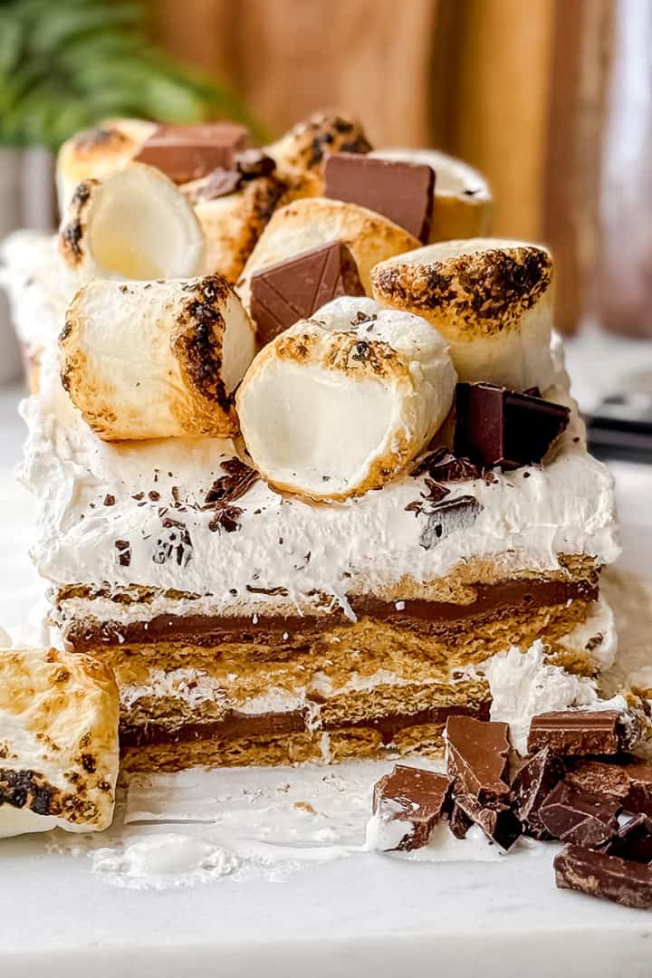 A no-bake s'mores icebox cake sits on a white surface, layered and topped with toasted marshmallows and chocolate pieces, with extra chocolate and marshmallows scattered in front.