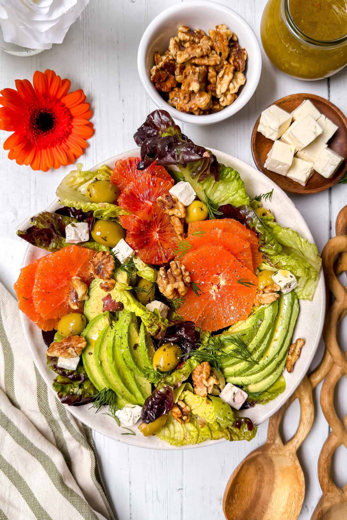 A colorful salad with avocado slices, orange and grapefruit segments, lettuce, green olives, walnuts, and feta cheese on a white plate, surrounded by a bowl of walnuts, feta cubes, dressing, and a red flower.