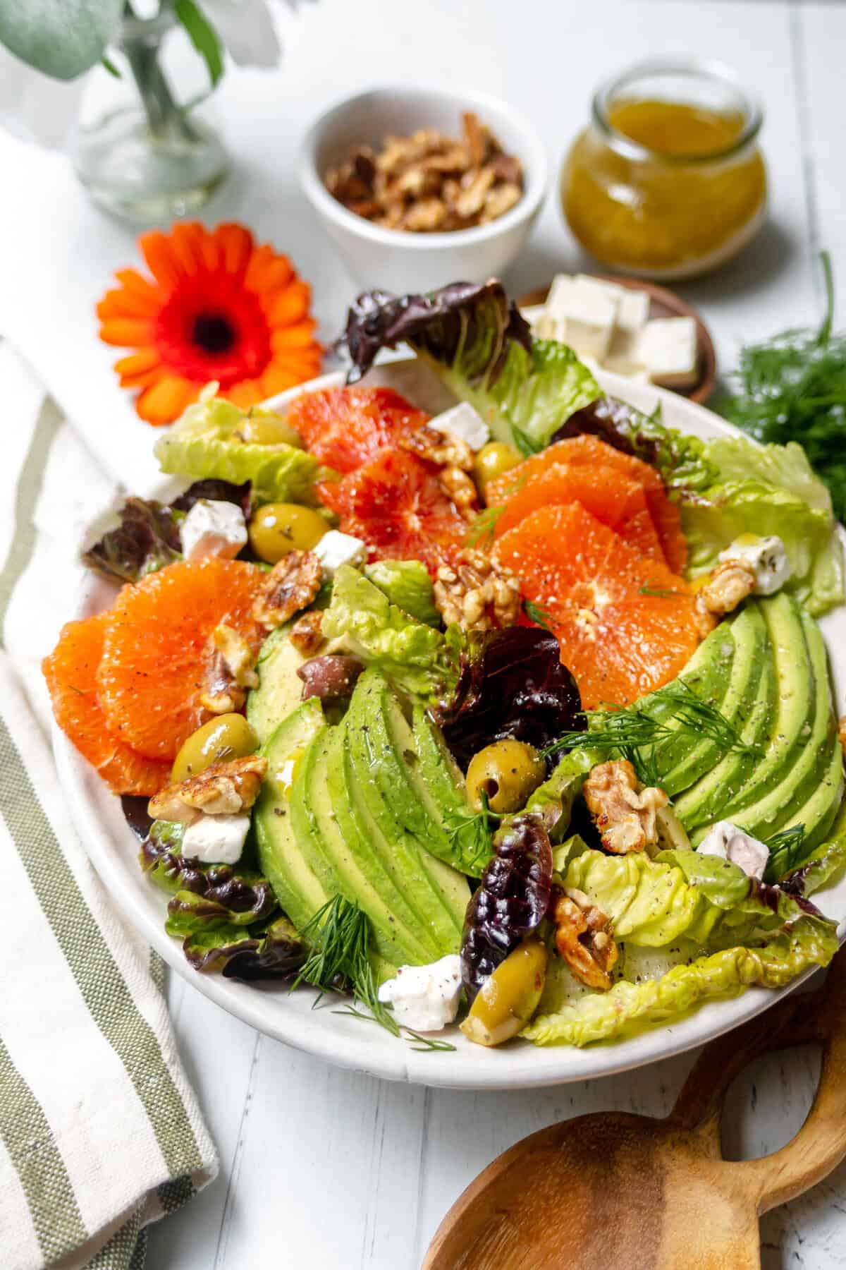 A fresh salad with avocado slices, orange and blood orange rounds, green olives, walnuts, mixed greens, feta cheese, and herbs on a white plate, with a jar of dressing, walnuts, and a flower in the background.