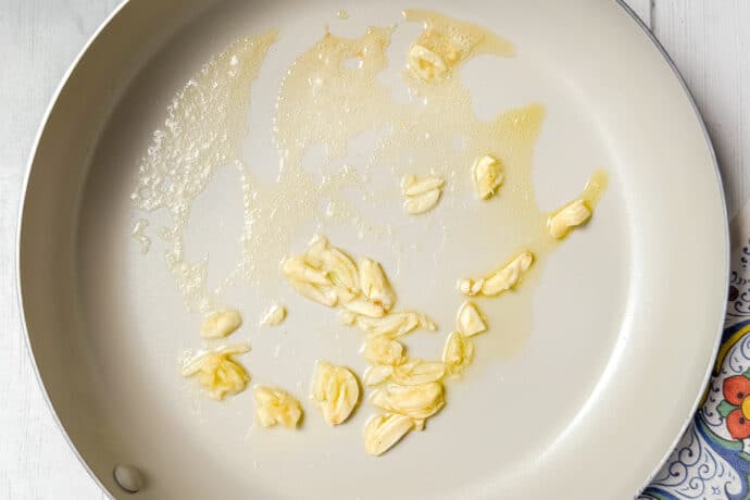 A frying pan on a white wooden surface contains sliced garlic sautéing in oil. A decorative cloth with a floral pattern and a white pitcher are partially visible beside the pan.
