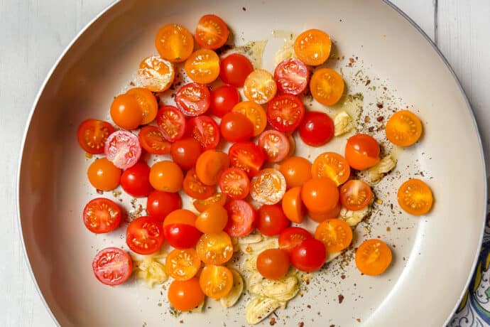 A skillet on a white surface contains halved red and yellow cherry tomatoes and sliced garlic, sprinkled with spices. A colorful patterned cloth is nearby, and part of a plant and pitcher are visible in the background.