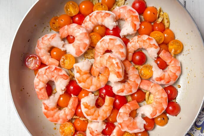 A skillet contains cooked shrimp, halved cherry tomatoes in red and yellow, and slices of garlic on a white wooden surface next to a colorful patterned cloth.