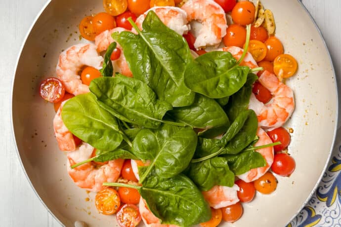 A skillet filled with cooked shrimp, halved cherry tomatoes, and fresh spinach leaves sits on a white wooden surface next to a patterned cloth.