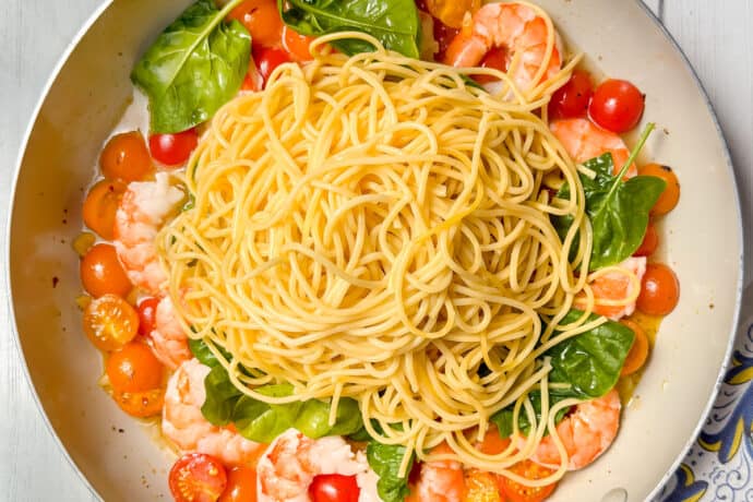A skillet filled with cooked spaghetti, shrimp, spinach, and cherry tomatoes sits on a white wooden table, surrounded by fresh greens and a colorful patterned napkin.