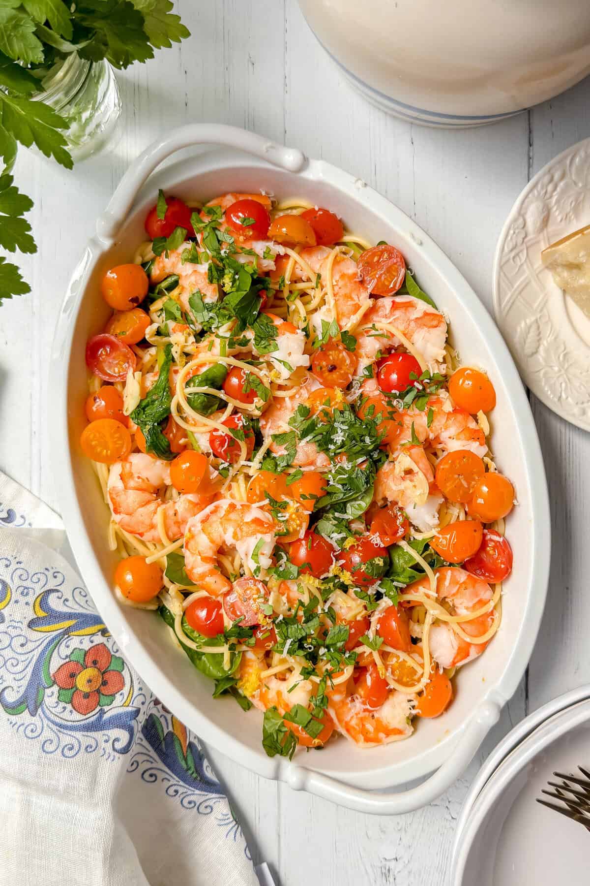 Oval white dish filled with spaghetti, shrimp, cherry tomatoes, and chopped parsley on a white wooden table, with a floral napkin, plate, and parsley garnish nearby.