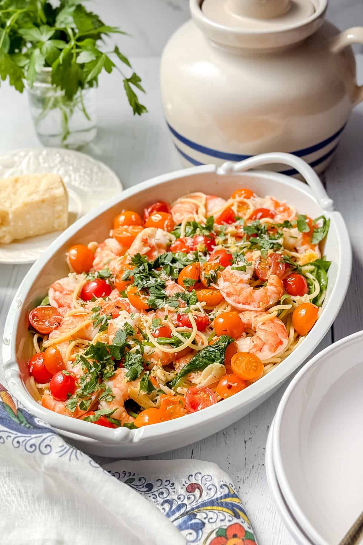 A white dish filled with spaghetti, shrimp, cherry tomatoes, spinach, and fresh parsley, set on a table with a block of cheese, a jar of parsley, and stacked plates nearby.