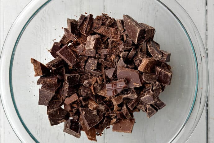 A clear glass bowl filled with chunks of dark chocolate sits on a white wooden surface, ready to be used in a delicious no-bake s'mores icebox cake.