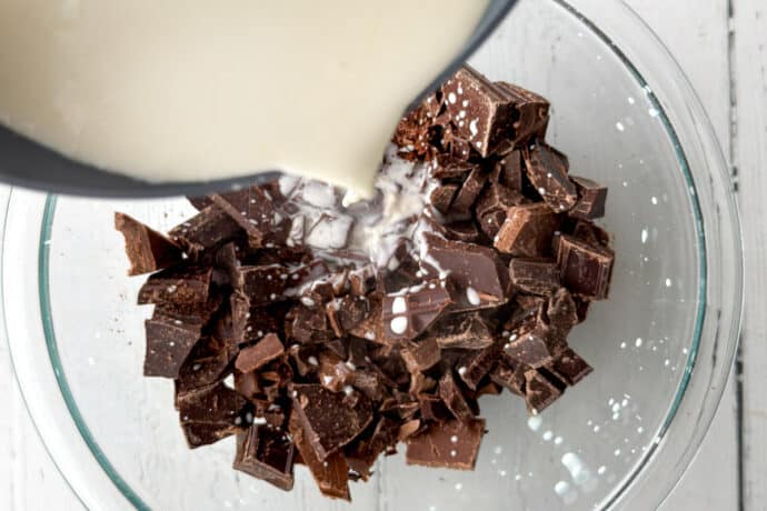 A bowl filled with chopped chocolate pieces as warm cream is poured over them, preparing to make a rich ganache for a no-bake s'mores icebox cake, on a white wooden surface.