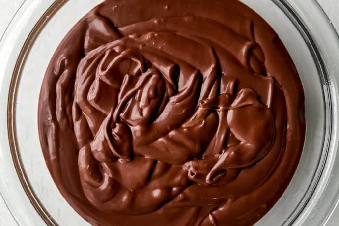 A glass bowl filled with smooth, swirled chocolate pudding—reminiscent of a no-bake s'mores icebox cake—sits on a white wooden surface, viewed from above.