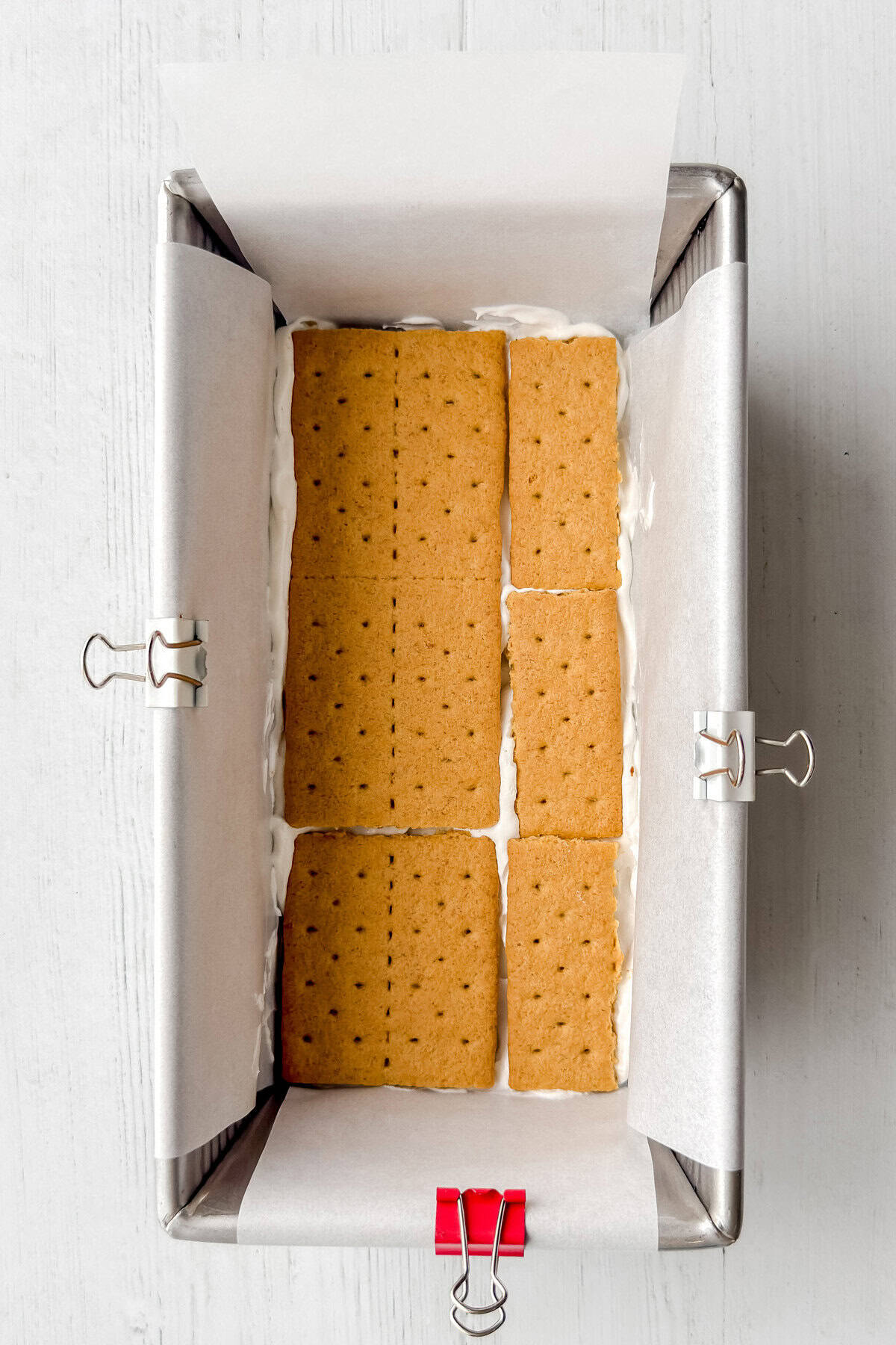 Overhead view of a loaf pan lined with parchment paper and metal binder clips, filled with a layer of graham crackers over a cream filling—perfect for assembling a no-bake s'mores icebox cake. The pan sits on a light-colored wooden surface.